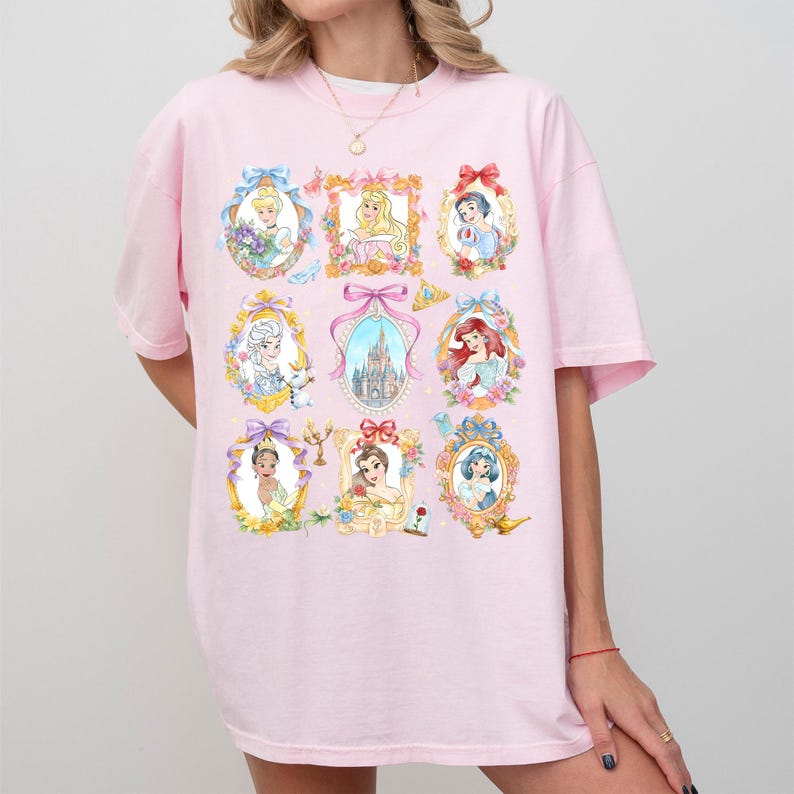 Pu&ograve; includere: T-shirt rosa chiaro con ritratti ad acquerello di principesse in cornici ornate. I ritratti sono circondati da motivi floreali e fiocchi. La maglietta ha maniche corte e scollo tondo.