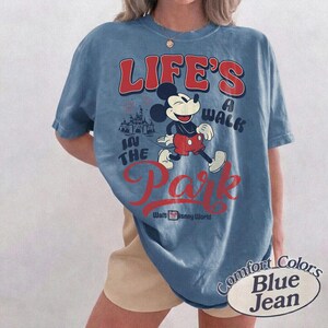 May include: Blue jean Comfort Colors t-shirt featuring Mickey Mouse walking in front of a castle with the text "LIFE'S A WALK IN THE PARK" in red. The shirt also says "Walt Disney World".