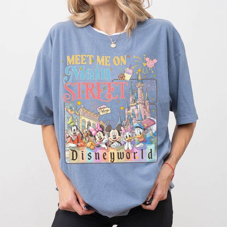 May include: A light blue t-shirt featuring a colorful graphic with the text "Meet Me On Main Street" and "Disneyworld." The graphic includes cartoon characters and a castle. The shirt has short sleeves and a relaxed fit.
