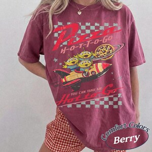 Pu&ograve; includere: T-shirt color bacca con grafica "Pizza H-O-T-T-O-Go" e un razzo che trasporta tre alieni verdi. Una pizza orbita un pianeta. La maglietta ha un aspetto vintage e usurato.