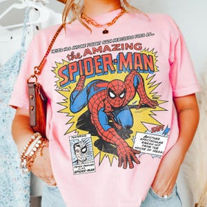 以下が含まれることがあります： スパイダーマンのコミック風グラフィックが特徴の薄いピンク色のTシャツ。「the AMAZING SPIDER-MAN」の文字とコミックの効果音が含まれています。Tシャツは薄いピンク色です。