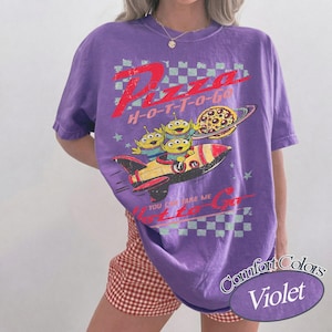 Pu&ograve; includere: Una maglietta Comfort Colors viola con una grafica di tre alieni verdi in un razzo, un pianeta pizza e la scritta "Pizza H-O-T-T-O-G-O". La maglietta ha un motivo a scacchi e la scritta "You can take me to go".