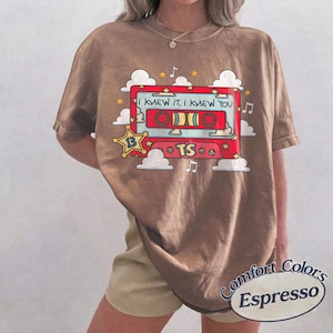 May include: A brown short-sleeve t-shirt featuring a graphic of a red cassette tape with the text I KNEW IT, I KNEW YOU and TS. The design includes white clouds, yellow stars, and musical notes surrounding the cassette tape illustration.