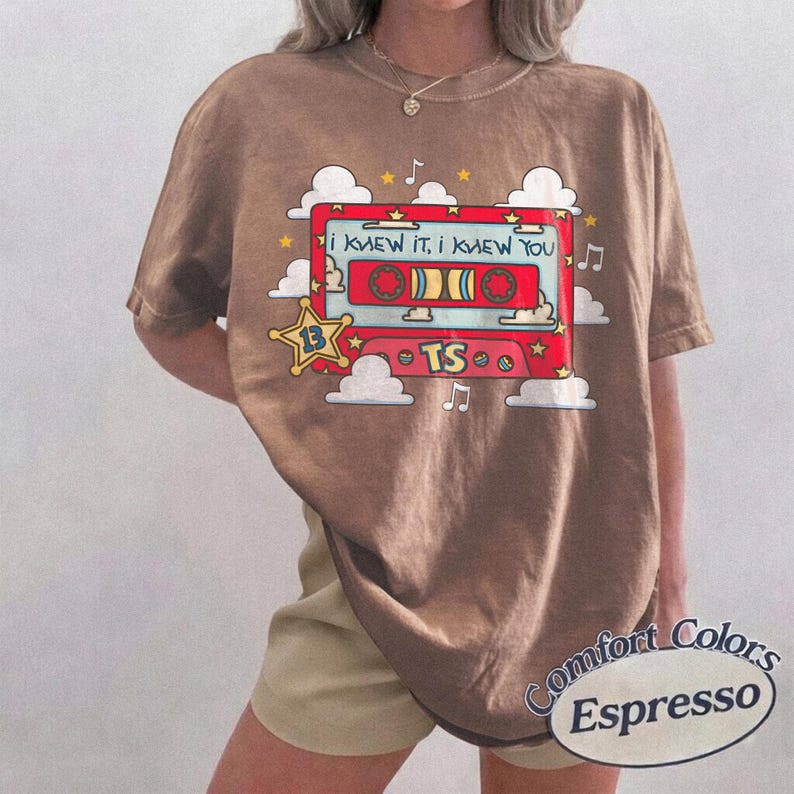 May include: A brown short-sleeve t-shirt featuring a graphic of a red cassette tape with the text I KNEW IT, I KNEW YOU and TS. The design includes white clouds, yellow stars, and musical notes surrounding the cassette tape illustration.