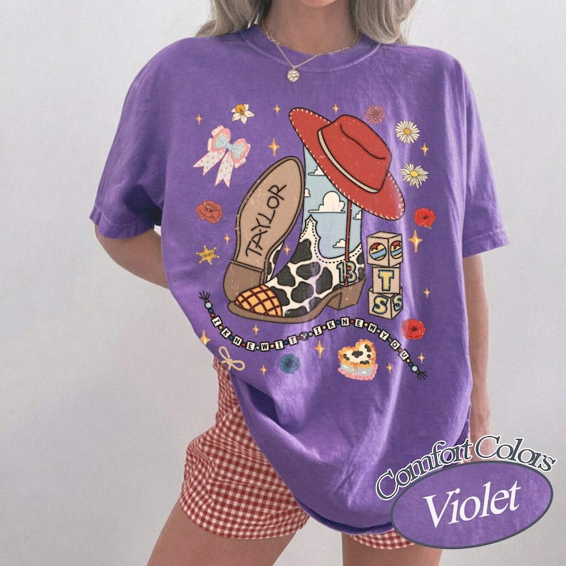 K&ouml;nnte beinhalten: Ein violettes Kurzarm-T-Shirt mit einem Grafikdruck, der einen Cowboystiefel mit Kuhfleckenmuster, einen roten Hut, Bl&ouml;cke mit den Buchstaben T, S und der Zahl 13, eine Schleife, Blumen sowie den Text Taylor und I KNEW IT I KNEW YOU zeigt.