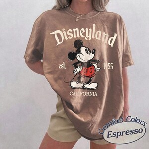 Może przedstawiać: Koszulka w kolorze espresso z grafiką Myszki Miki i napisem "Disneyland" w kolorze białym. Koszulka zawiera r&oacute;wnież tekst "est. 1955" i "CALIFORNIA". Koszulka marki Comfort Colors.