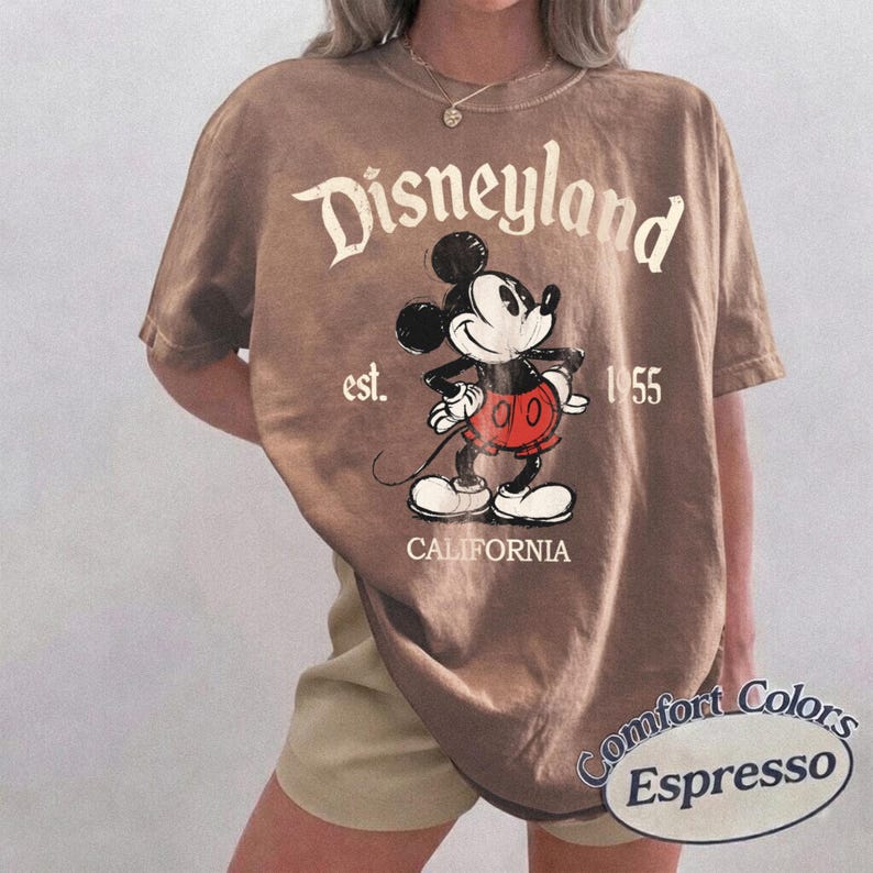 Może przedstawiać: Koszulka w kolorze espresso z grafiką Myszki Miki i napisem "Disneyland" w kolorze białym. Koszulka zawiera r&oacute;wnież tekst "est. 1955" i "CALIFORNIA". Koszulka marki Comfort Colors.