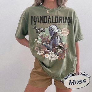 Może przedstawiać: Oliwkowa koszulka z nadrukiem The Mandalorian i Grogu otoczonych kwiatami. Tekst "THE MANDALORIAN" i "THIS IS THE WAY" jest wydrukowany nad obrazem. Koszulka jest oznaczona jako "Comfort Colors Moss".