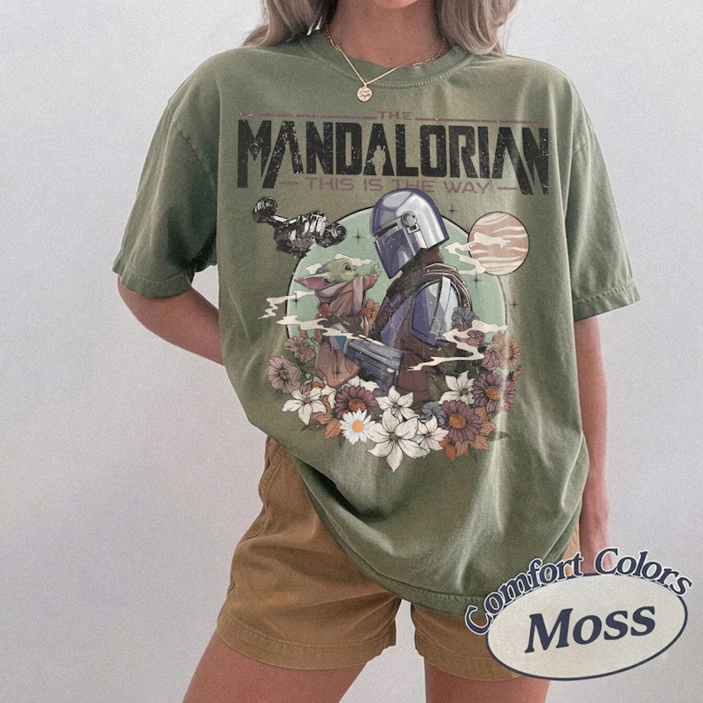 Może przedstawiać: Oliwkowa koszulka z nadrukiem The Mandalorian i Grogu otoczonych kwiatami. Tekst "THE MANDALORIAN" i "THIS IS THE WAY" jest wydrukowany nad obrazem. Koszulka jest oznaczona jako "Comfort Colors Moss".
