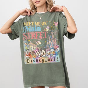 May include: Olive green t-shirt with a graphic featuring Disney characters and the text "Meet Me On Main Street Disneyworld." The design includes a castle, fireworks, and various cartoon characters. The shirt has short sleeves and a relaxed fit.