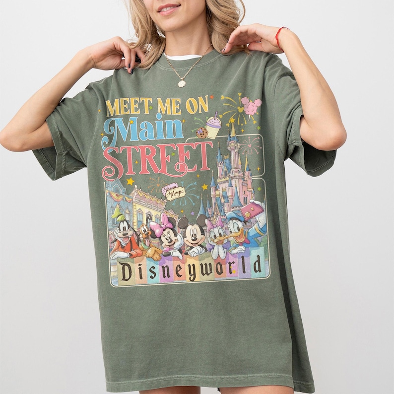 May include: Olive green t-shirt with a graphic featuring Disney characters and the text "Meet Me On Main Street Disneyworld." The design includes a castle, fireworks, and various cartoon characters. The shirt has short sleeves and a relaxed fit.