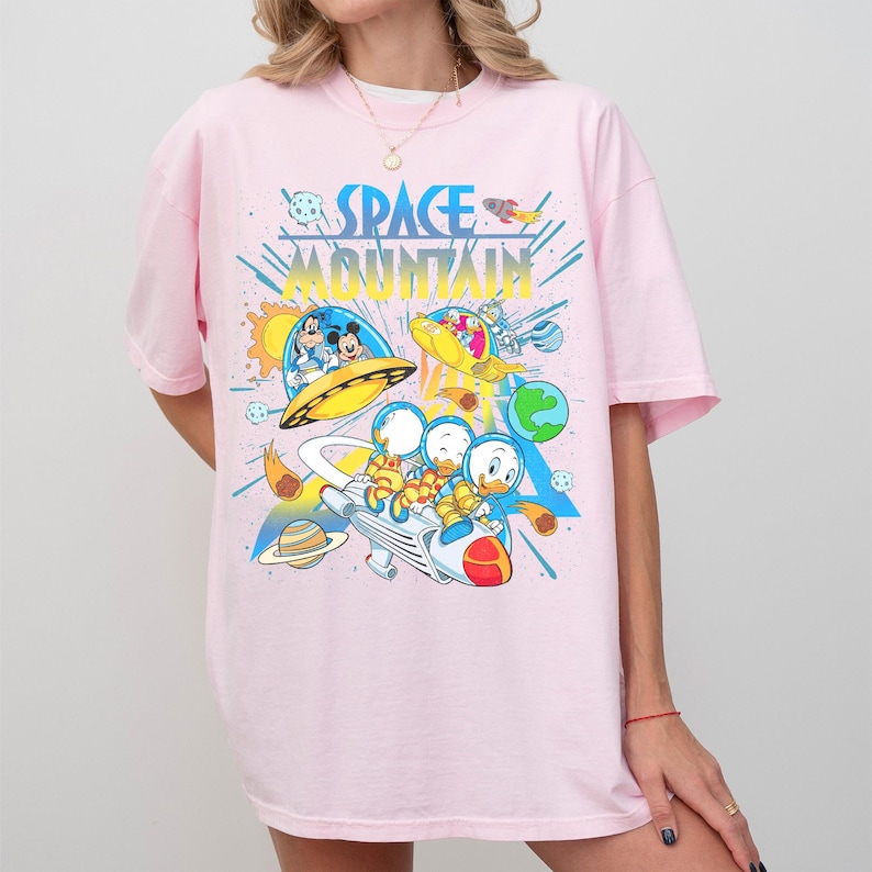 Pu&ograve; includere: T-shirt rosa chiaro con un design grafico colorato. Il design presenta le parole "SPACE MOUNTAIN" in blu e giallo, insieme a personaggi dei cartoni animati in veicoli spaziali, con pianeti e stelle.