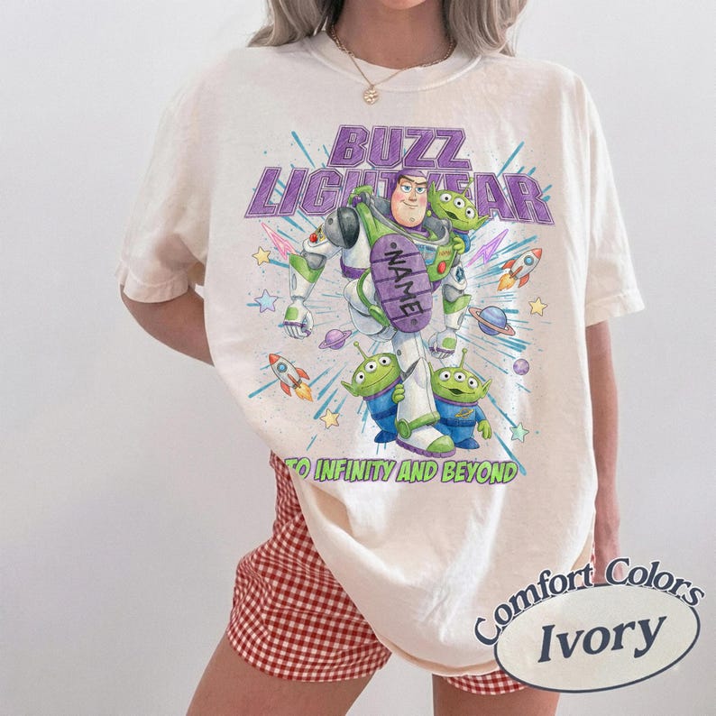 May include: An ivory t-shirt featuring a graphic of Buzz Lightyear and three green aliens from Toy Story. The text reads BUZZ LIGHTYEAR and TO INFINITY AND BEYOND, with a purple oval containing the word NAME. Colorful rockets and stars surround them.