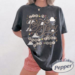 May include: A charcoal gray short-sleeve t-shirt featuring a graphic print with the text "ITS A TAY STORY BABY JUST SAY YES". The design includes rope-style lettering, musical notes, stars, and cloud shapes in white, yellow, and blue colors.