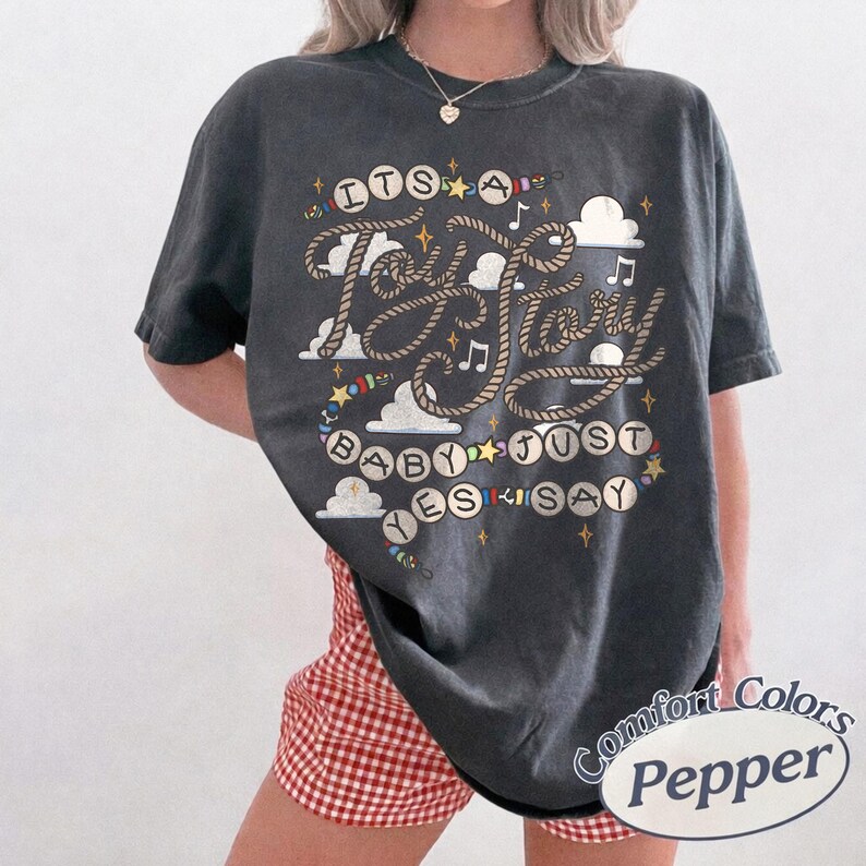 May include: A charcoal gray short-sleeve t-shirt featuring a graphic print with the text "ITS A TAY STORY BABY JUST SAY YES". The design includes rope-style lettering, musical notes, stars, and cloud shapes in white, yellow, and blue colors.
