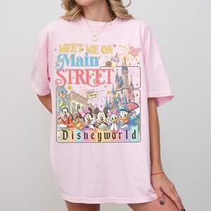 May include: A light pink t-shirt featuring a colorful graphic with the text "Meet Me On Main Street Disneyworld." The graphic includes cartoon characters and a castle. The shirt has short sleeves and a crew neck.