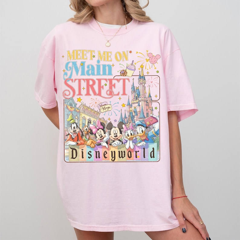 May include: A light pink t-shirt featuring a colorful graphic with the text "Meet Me On Main Street Disneyworld." The graphic includes cartoon characters and a castle. The shirt has short sleeves and a crew neck.