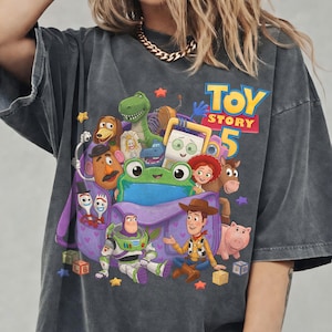 May include: A charcoal gray short-sleeve t-shirt featuring a colorful graphic print of various Toy Story characters, including Woody, Buzz Lightyear, and a green frog, centered around a purple bag with the text Toy Story 5 in blue and yellow.