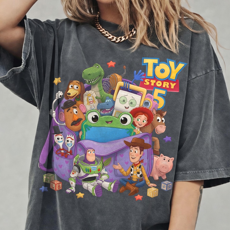May include: A charcoal gray short-sleeve t-shirt featuring a colorful graphic print of various Toy Story characters, including Woody, Buzz Lightyear, and a green frog, centered around a purple bag with the text Toy Story 5 in blue and yellow.