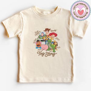 May include: A cream-colored short-sleeve t-shirt featuring a graphic print of Toy Story characters including Woody, Buzz Lightyear, Jessie, Mr. Potato Head, Hamm, Rex, and Slinky Dog, with their names written in cursive and the Toy Story logo below.
