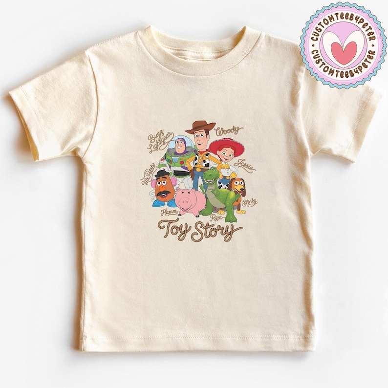 May include: A cream-colored short-sleeve t-shirt featuring a graphic print of Toy Story characters including Woody, Buzz Lightyear, Jessie, Mr. Potato Head, Hamm, Rex, and Slinky Dog, with their names written in cursive and the Toy Story logo below.