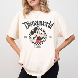 May include: Cream-colored t-shirt featuring a vintage Disney World graphic. The design includes Mickey Mouse, the words "Disneyworld," "Florida," and the year "1971." The graphic is in black and red, with a checkered border around Mickey.