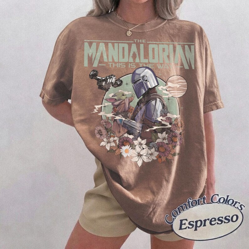 Może przedstawiać: Brązowy t-shirt w kolorze espresso z grafiką z "The Mandalorian". Projekt zawiera napis "THE MANDALORIAN - THIS IS THE WAY" nad ilustracją Mandalorianina i Grogu otoczonych kwiatami. Koszulka jest z Comfort Colors.