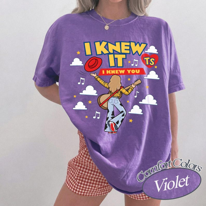 May include: A violet short-sleeve t-shirt featuring a graphic of a person playing a guitar with the text I KNEW IT and I KNEW YOU. The design includes a red hat, a red heart, white clouds, yellow stars, and musical notes. The shirt has a crew neckline.