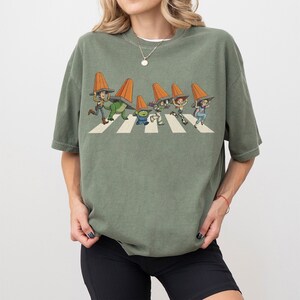 Puede incluir: Camiseta verde salvia con un gr&aacute;fico de personajes de dibujos animados cruzando una calle, cada uno con un cono de tr&aacute;fico naranja. Los personajes caminan sobre un paso de cebra blanco. La camiseta es de corte holgado.