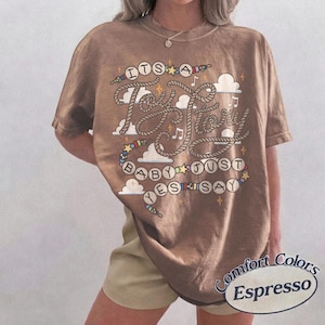 May include: An espresso brown short-sleeve t-shirt featuring a graphic print with the text "ITS A Toy Story BABY JUST YES SAY". The design includes rope-style lettering, colorful beads, white clouds, and small yellow stars scattered across the front.