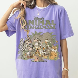 Puede incluir: Una camiseta morada con el texto "Disney's Animal Kingdom" y un gr&aacute;fico de varios personajes y animales de Disney, incluyendo Mickey Mouse, Donald Duck y un tigre. La camiseta tiene mangas cortas y un corte holgado.