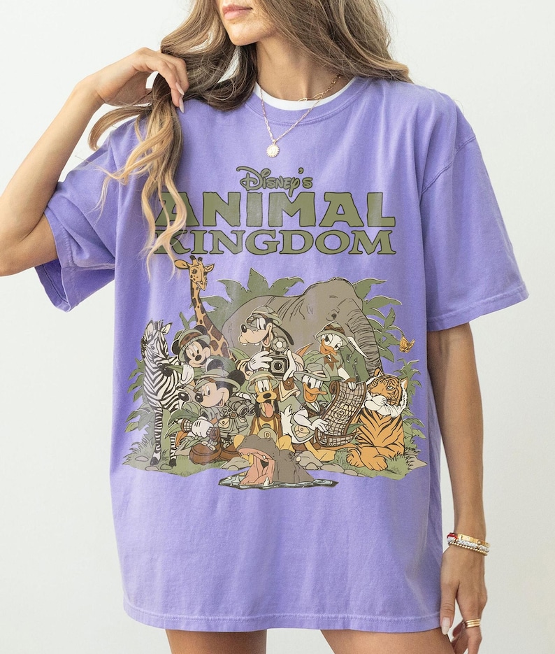 Puede incluir: Una camiseta morada con el texto "Disney's Animal Kingdom" y un gr&aacute;fico de varios personajes y animales de Disney, incluyendo Mickey Mouse, Donald Duck y un tigre. La camiseta tiene mangas cortas y un corte holgado.