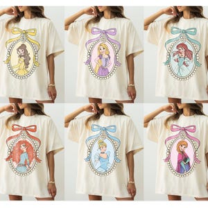 May include: Cream-colored oversized t-shirts featuring cartoon portraits of Disney princesses within ornate oval frames. Each frame is topped with a decorative bow in various colors, including yellow, purple, teal, red, blue, and pink.