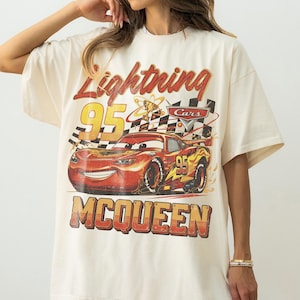 May include: Cream-colored t-shirt featuring a graphic of Lightning McQueen from the Cars movie. The design includes the text "Lightning McQueen 95" and "McQueen" in a vintage style, with a checkered flag and trophy.