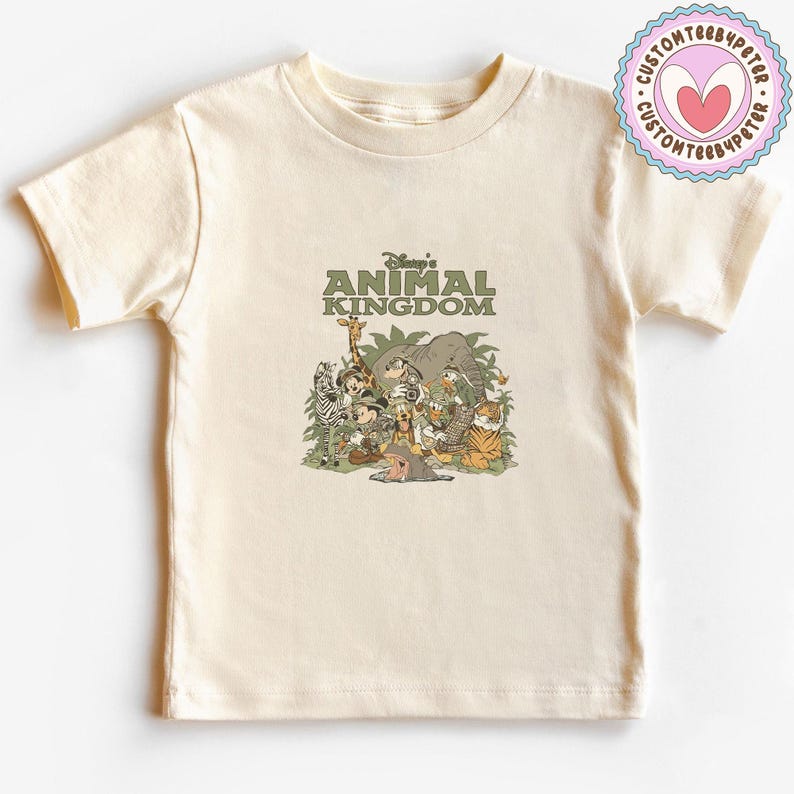Puede incluir: Camiseta color crema con el texto "Disney's Animal Kingdom" sobre un gr&aacute;fico de varios animales de dibujos animados, incluyendo una cebra, una jirafa, un elefante y un tigre. El dise&ntilde;o es en tonos verdes, marrones y beige.