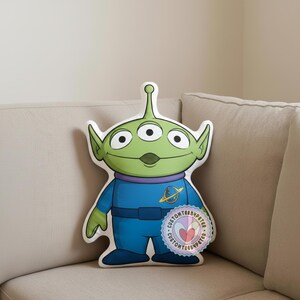 Peut inclure: Un coussin en forme de l'Alien Vert de Toy Story. Le coussin pr&eacute;sente une t&ecirc;te verte avec trois yeux, une combinaison bleue et un col violet. Un autocollant circulaire avec du texte est visible en bas &agrave; droite.