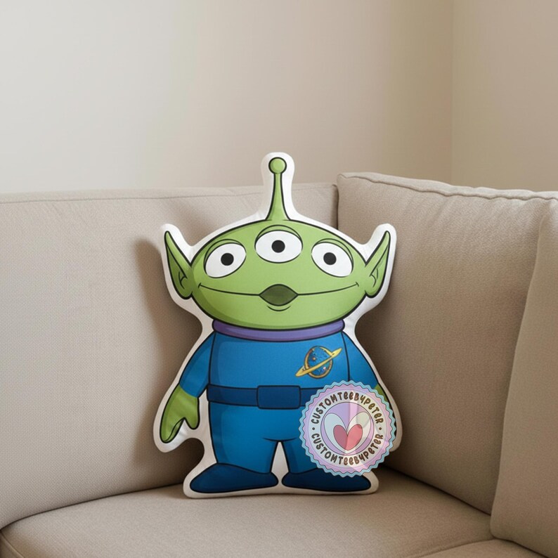 Peut inclure: Un coussin en forme de l'Alien Vert de Toy Story. Le coussin pr&eacute;sente une t&ecirc;te verte avec trois yeux, une combinaison bleue et un col violet. Un autocollant circulaire avec du texte est visible en bas &agrave; droite.