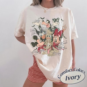 May include: Ivory Comfort Colors t-shirt with a Halloween-themed Disney character graphic. The design includes Mickey Mouse, Minnie Mouse, Pluto, Daisy Duck, and Goofy in costumes, with their signatures. The shirt is worn with red and white checked shorts.