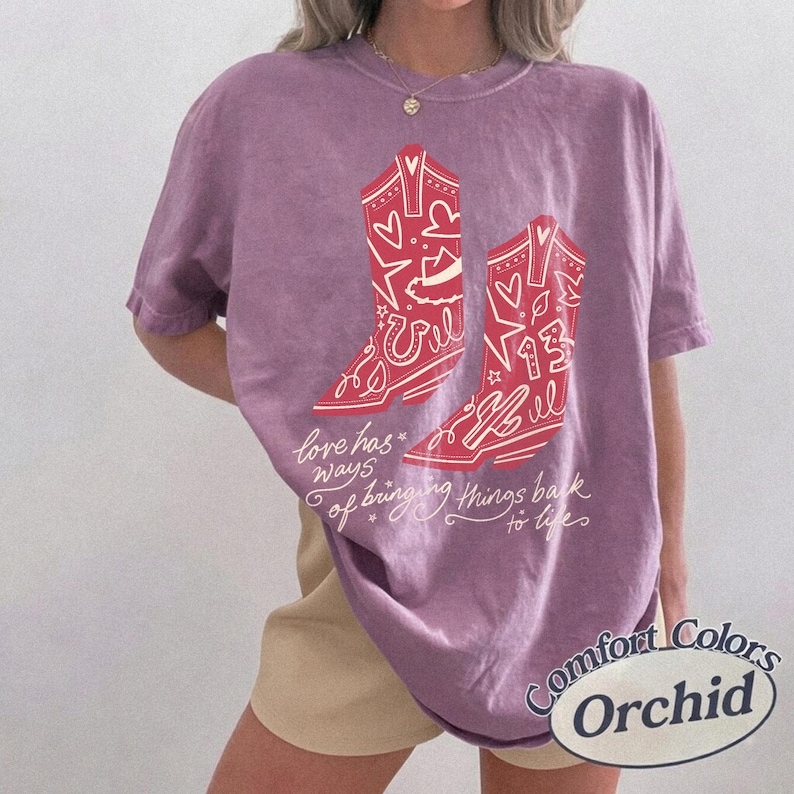 May include: A purple short-sleeve t-shirt featuring a graphic of two red cowboy boots decorated with hearts, stars, and the number 13. The text printed on the front reads, love has ways of bringing things back to life, in a white cursive script.