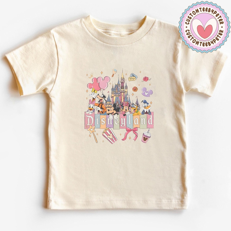 May include: Cream-colored t-shirt featuring a colorful Disneyland graphic. The design includes a castle, Disney characters, and the word "Disneyland" in pastel letters. A pink and purple logo is on the upper right.