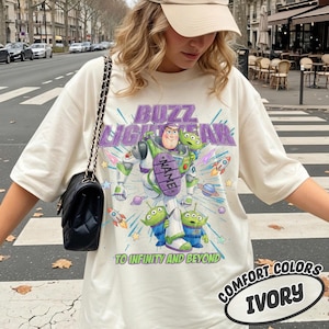 May include: An ivory t-shirt featuring a graphic of Buzz Lightyear and two green aliens. The text reads BUZZ LIGHTYEAR and TO INFINITY AND BEYOND, with a purple oval containing the word NAME. A black quilted shoulder bag with a chain strap is worn.