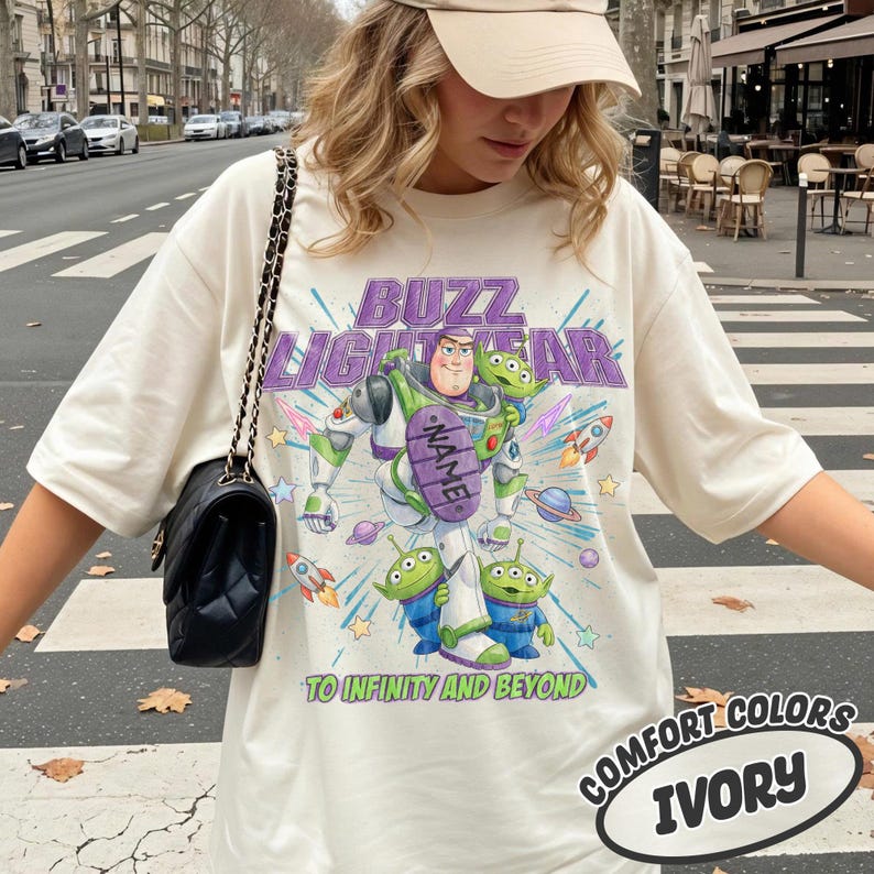 May include: An ivory t-shirt featuring a graphic of Buzz Lightyear and two green aliens. The text reads BUZZ LIGHTYEAR and TO INFINITY AND BEYOND, with a purple oval containing the word NAME. A black quilted shoulder bag with a chain strap is worn.