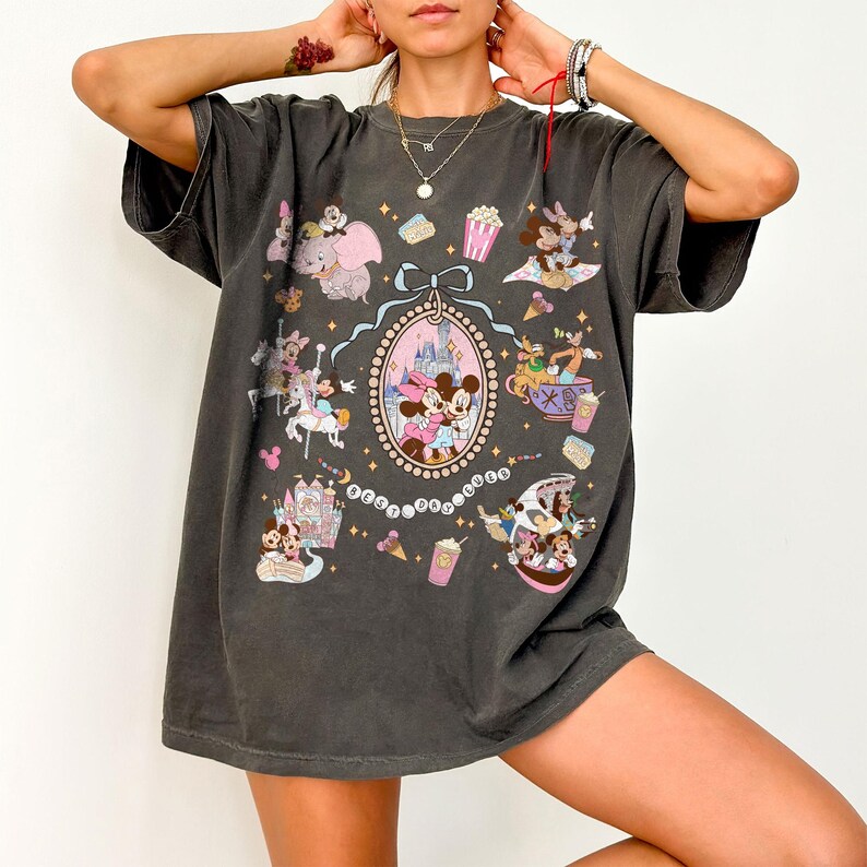 May include: Dark gray t-shirt featuring a colorful graphic of Disney characters and attractions. The design includes Mickey and Minnie Mouse, Dumbo, and a castle, surrounded by various themed elements. The shirt has a relaxed fit and short sleeves.