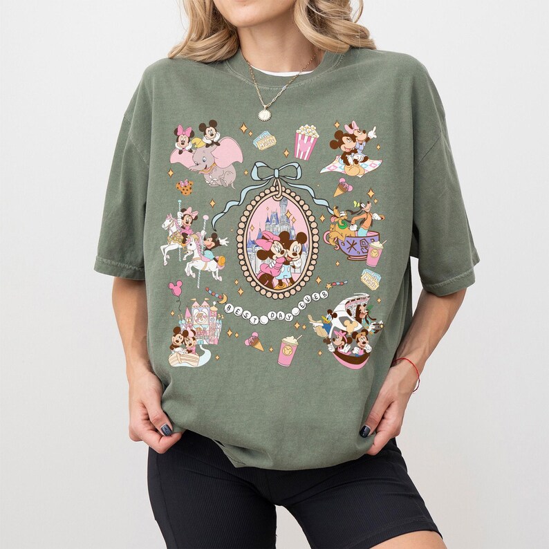 May include: Olive green t-shirt featuring a colorful graphic of Disney characters and icons. The design includes Mickey and Minnie Mouse, a castle, and other theme park elements. The shirt has a relaxed fit and short sleeves.