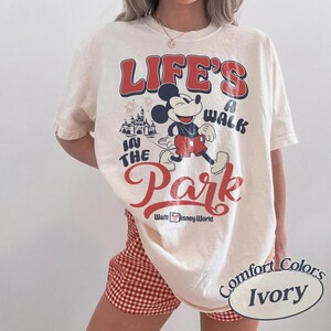 May include: Ivory-coloured t-shirt featuring Mickey Mouse walking in front of a castle, with the text "LIFE'S A WALK IN THE PARK" in red and black. The shirt is from Walt Disney World.