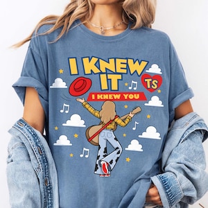 May include: A blue t-shirt featuring a graphic of a person with a guitar, a red hat, and clouds. The text reads I KNEW IT, I KNEW YOU, and TS inside a red heart. The person wears a yellow plaid shirt and cow-print trousers with red shoes.