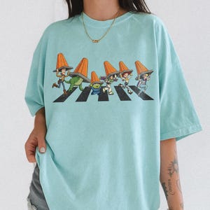 Puede incluir: Camiseta azul claro con un dise&ntilde;o de personajes de Toy Story cruzando un paso de cebra, cada uno con un cono de tr&aacute;fico naranja. El dise&ntilde;o es una parodia de la ic&oacute;nica portada del &aacute;lbum Abbey Road. La camiseta es de corte holgado.