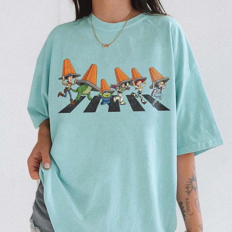 Puede incluir: Camiseta azul claro con un dise&ntilde;o de personajes de Toy Story cruzando un paso de cebra, cada uno con un cono de tr&aacute;fico naranja. El dise&ntilde;o es una parodia de la ic&oacute;nica portada del &aacute;lbum Abbey Road. La camiseta es de corte holgado.