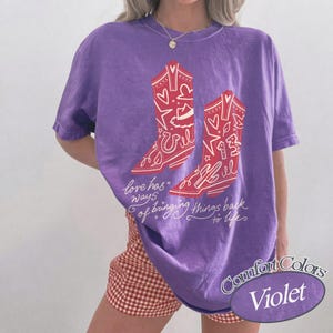 May include: A violet short-sleeved t-shirt featuring a graphic of two red cowboy boots decorated with hearts, stars, and the number 13. Below the boots, the text reads, "love has ways of bringing things back to life" in white cursive script.