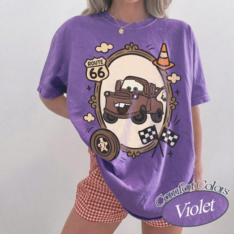 May include: A violet Comfort Colors t-shirt featuring a cartoon image of Mater from the movie Cars, framed by a decorative border with Route 66 signage, a traffic cone, and checkered flags. The shirt has a relaxed fit.